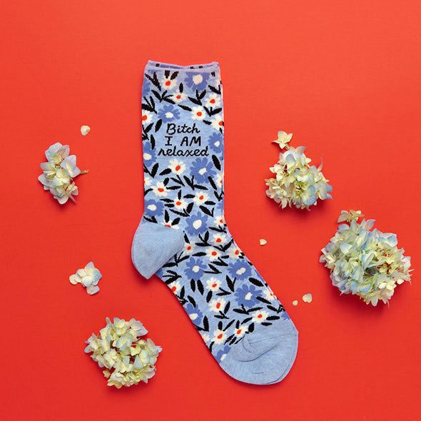 Funny floral socks for women that say, “Bitch, I AM relaxed” laying flat next to flowers on a red background
