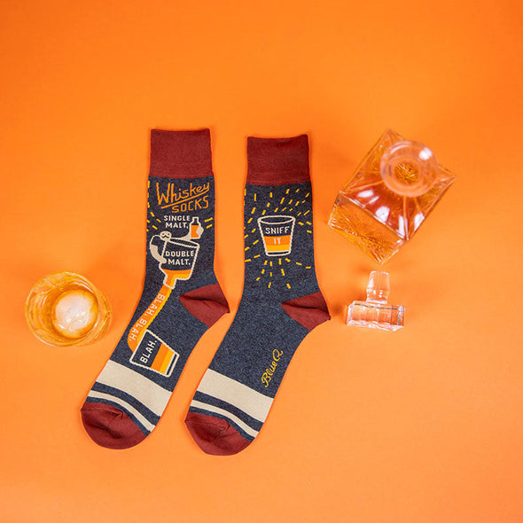 whiskey with ice in a glass next to funny men’s drinking socks that say “whiskey socks,” and “single malt, double malt, blah blah blah.”