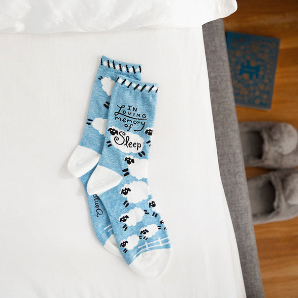 fun women's socks with sheep that say "in loving memory of sleep" lying on top of a bed