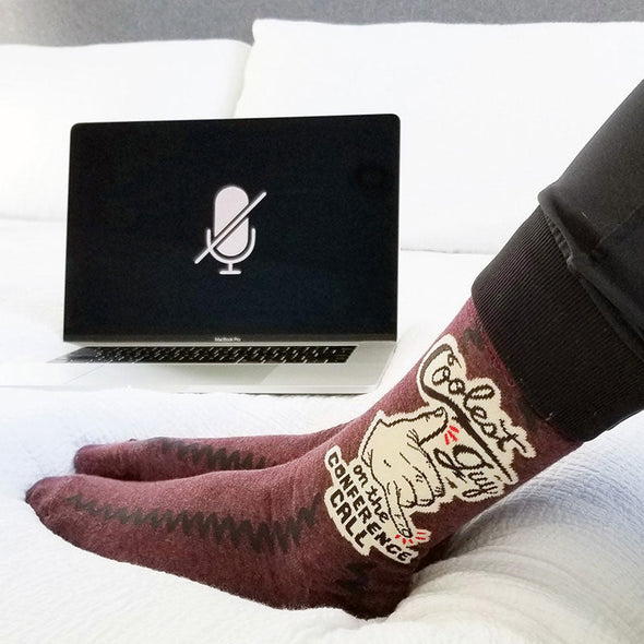 A man wearing The Coolest Guy on The Conference call socks sitting on a bed with laptop.