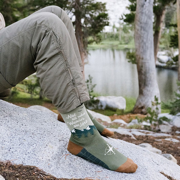 A man wearing sitting on a rock next to a lake wearing nature socks that say 'Not all who wander are lost'