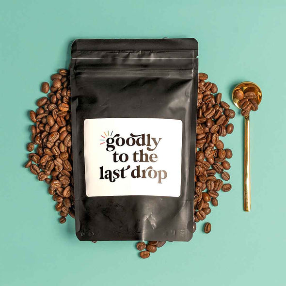 A bag of Goodly to the Last Drop coffee blend with gold spoon full of coffee beans