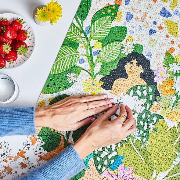 Hands putting together a puzzle of a woman in a bathtub surrounded by plants and flowers