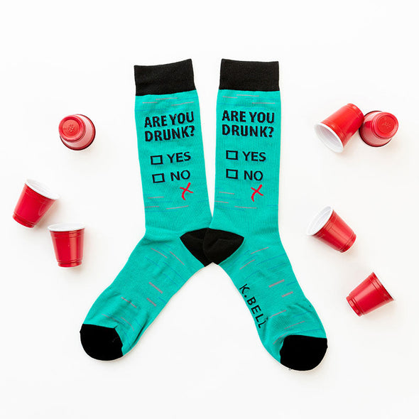 Funny men’s socks that say “Are you drunk?” laying flat with mini red solo cups scattered around the socks