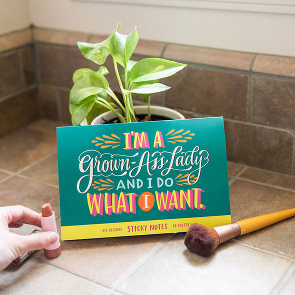 A funny pack of sticky notes that says, "I'm a grown-ass lady and I do what I want" sitting in between lipstick and a makeup brush