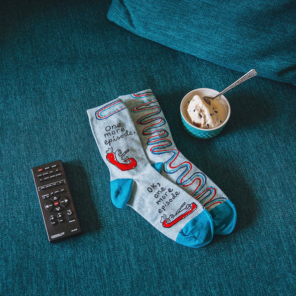 Funny "one more episode" socks for women laying flat next to a TV remote and a cup of ice cream