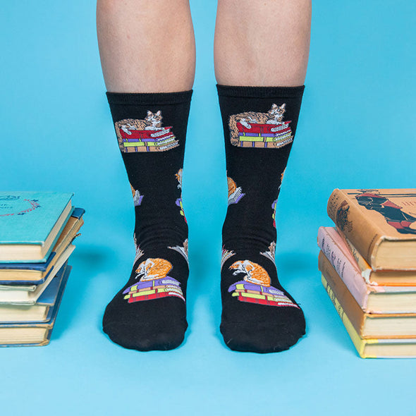 woman standing next to a stack of books and wearing socks with a pattern of cats sitting on books